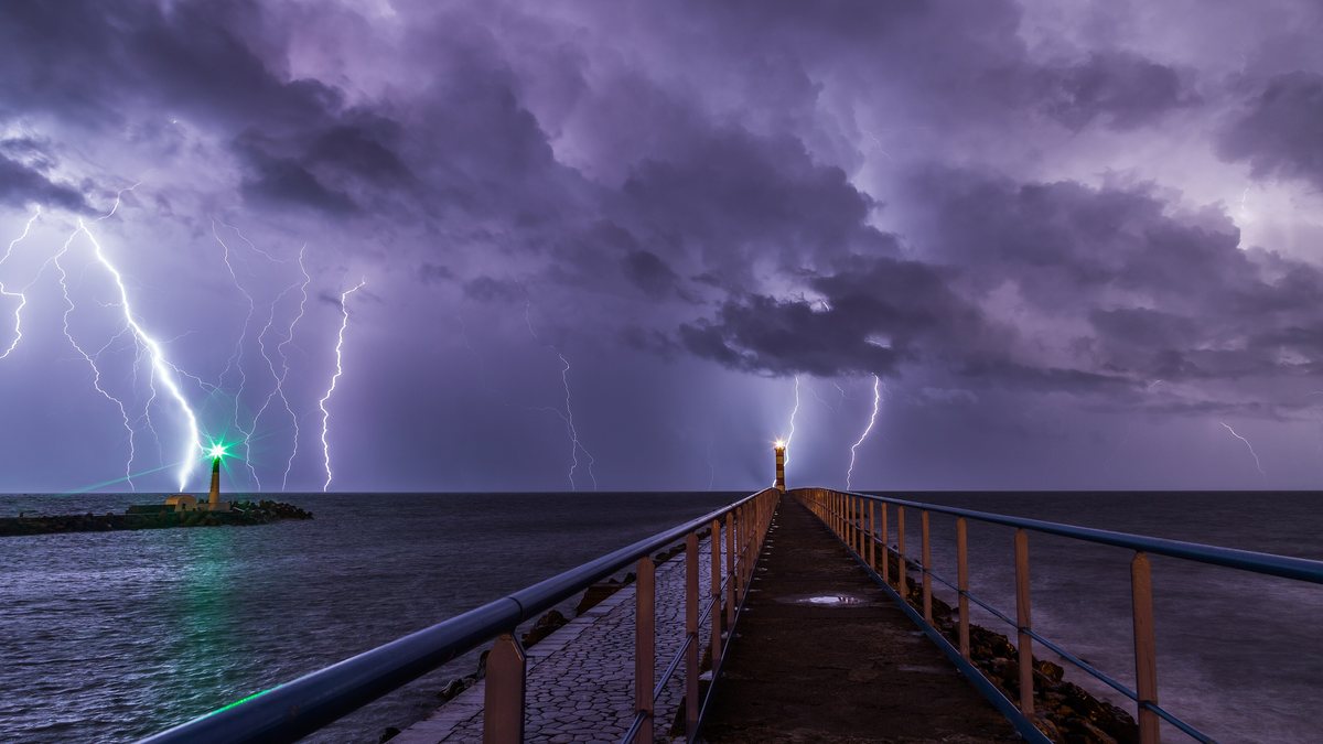 A lighthouse at the end of a jetty during a night-time lightning storm